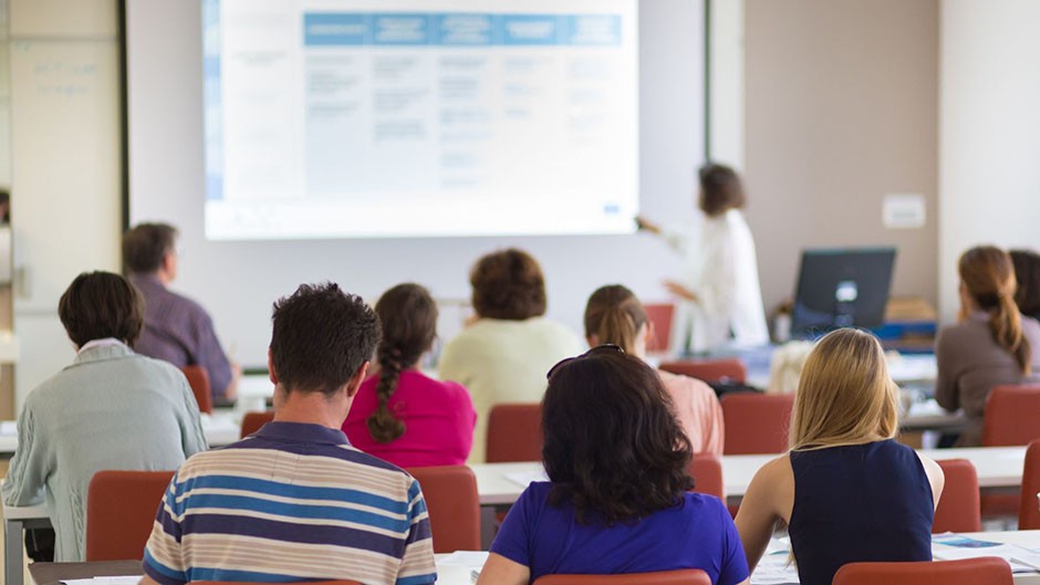 An instructor is teaching a course to students in a lecture hall using a screen and projector.