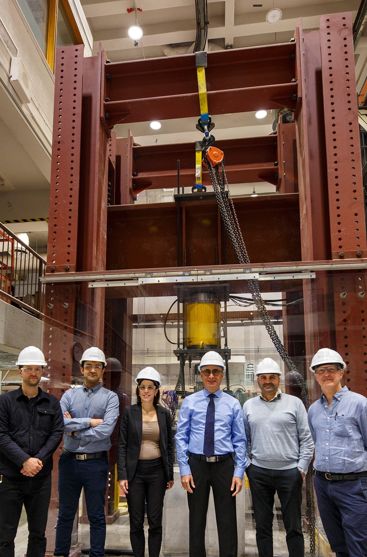 A group of six people in hard hats stands in a building next to a tall red metal structure.