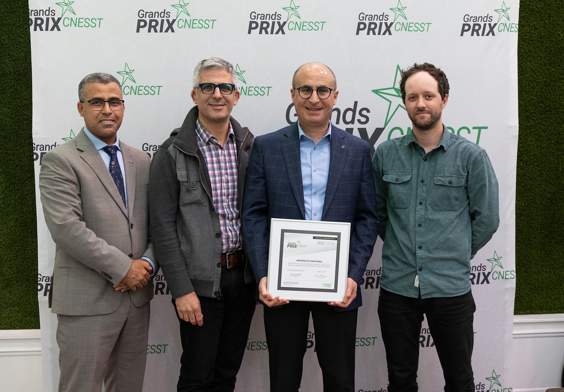 Four men stand side-by-side in front of a "Grands Prix CNESST" backdrop. One of them is holding a framed certificate.