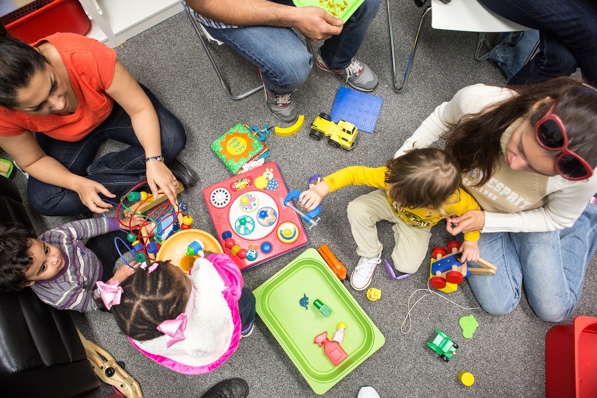 Two women sitting on a carpeted floor with three toddlers playing with toys. 