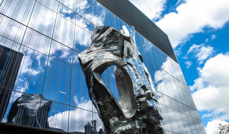 Grande sculpture en métal devant un immeuble aux fenêtres miroir.