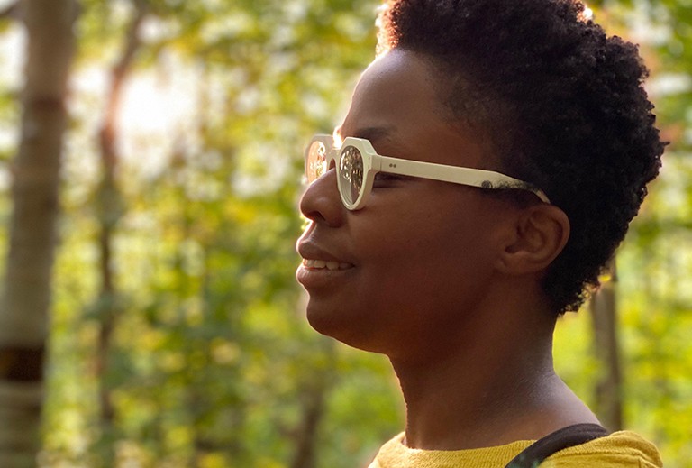 Side profile of young, smiling Black woman, with a yellow top and white-rimmed glasses.