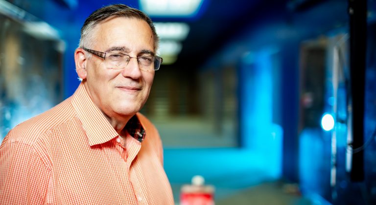 Older smiling gentleman with short grey hair and glasses and an orange shirt.