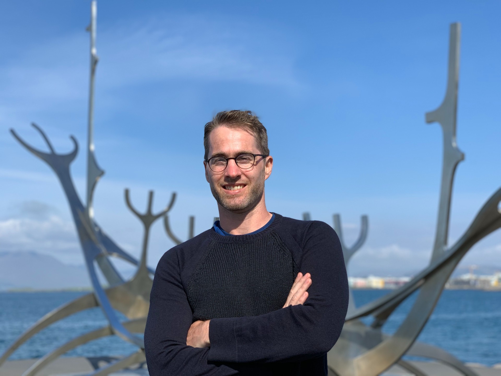 Man in black sweater standing by a river with a city in the background