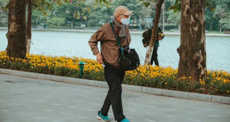 An older gentleman in a baseball cap and a mask walking along a boulevard.