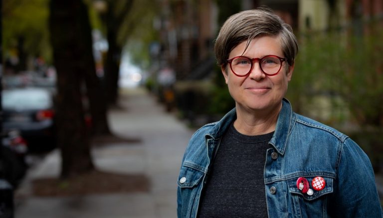 Woman with short hair, glasses and jean jacket standing on a sidewalk outside