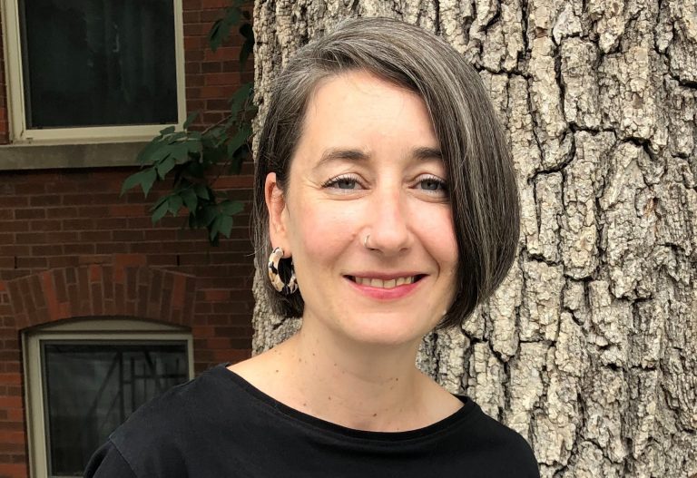 Femme souriante aux cheveux courts portant une chemise noire debout devant un tronc d'arbre et un bâtiment en briques rouges.