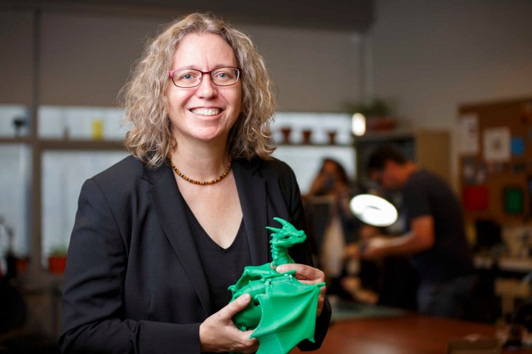 Femme souriante aux cheveux blonds bouclés mi-longs portant un blazer et une chemise noirs, tenant un dragon en plastique vert.