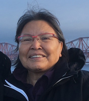Smiling Indigenous woman with red-rimmed glasses and wearing an outdoor jacket.