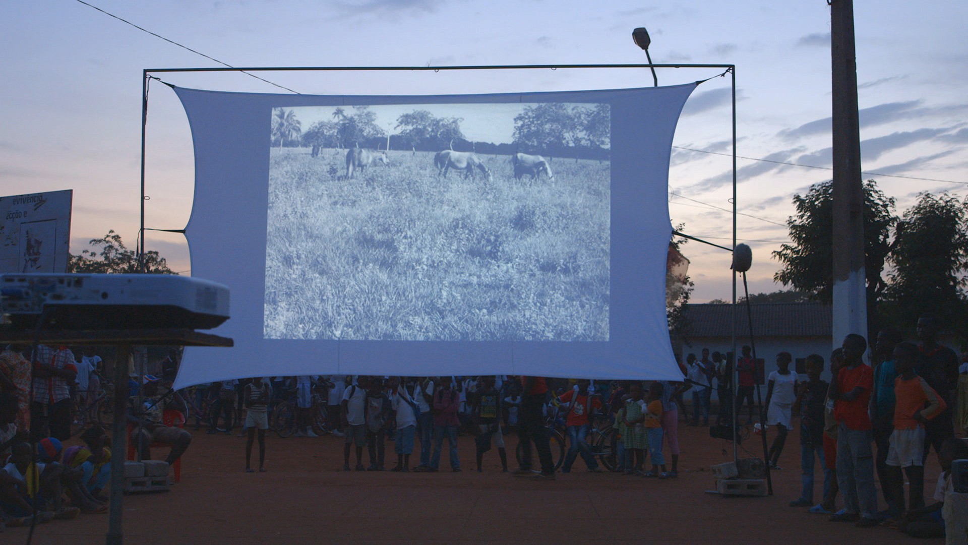 Spell Reel, 2017, par Filipa César | Image fournie par l'artiste Une grande feuille de projecteur tendue dans un décor extérieur avec des personnes debout autour, et représentant, en noir et blanc, des chevaux dans l'herbe longue.