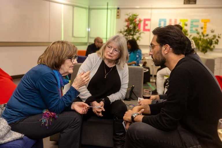 Trois femmes au premier plan se parlent lors d'un rassemblement de nombreuses personnes dans un intérieur.