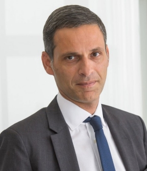 Man with greying close-cropped hair, wearing a charcoal grey suit and blue tie.