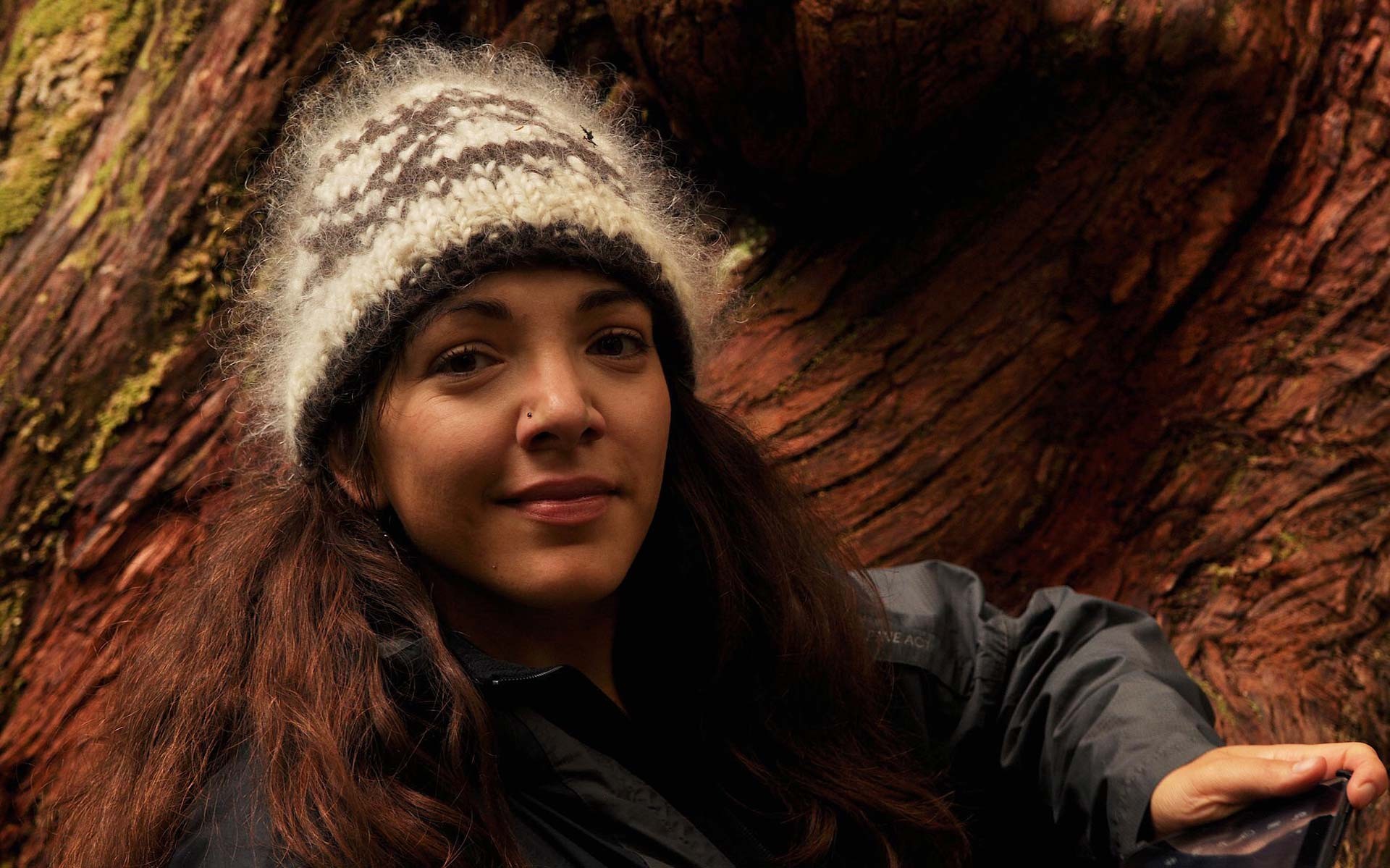 Une jeune femme souriante de face sur le tronc d'un énorme arbre, portant un bonnet de laine et une veste noire.