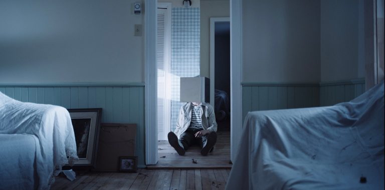 Un salon avec des meubles drapés et, dans l'embrasure de la porte, une personne assise sur le sol avec une sorte de boîte autour de la tête qui émet de la lumière.