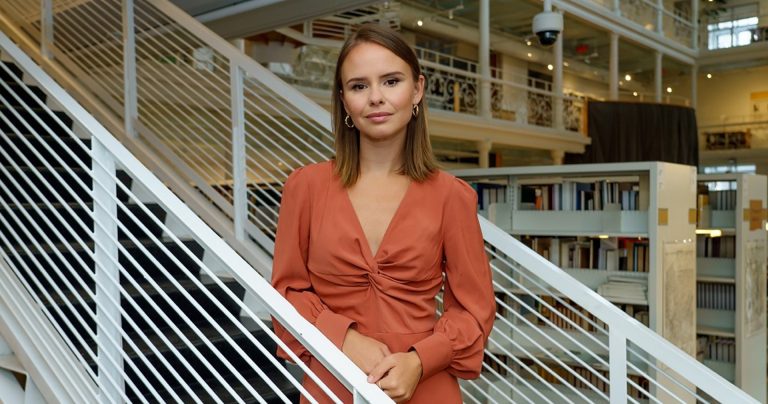 Femme aux longs cheveux blonds foncés, vêtue d'une robe orange et debout dans les escaliers.