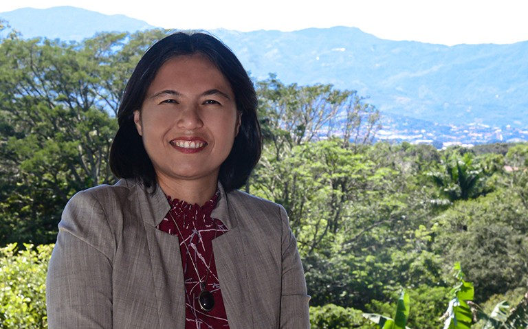 Femme souriante aux épaules foncées, vêtue d'une chemise rouge foncé et d'un blazer beige, debout en plein air, avec des arbres et des montagnes derrière elle.