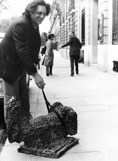 Homme souriant aux cheveux bruns mi-longs, vêtu d'un blazer et d'un pantalon et traînant une sorte de sculpture sur le trottoir.