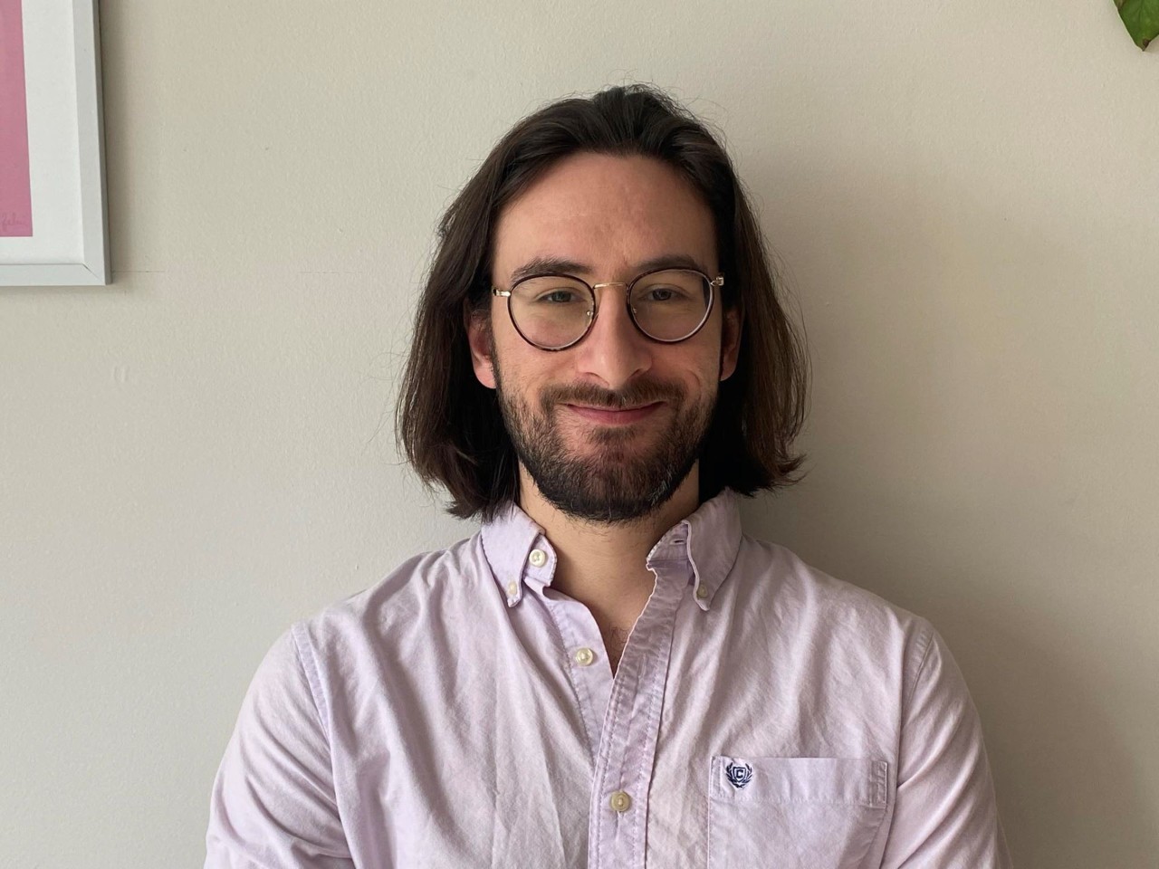 Un homme aux cheveux bruns mi-longs, portant des lunettes, une courte barbe et une chemise blanche se tient debout sur un fond blanc.