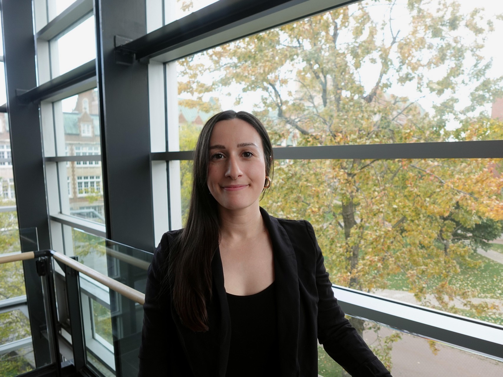 Stephanie Gumuchian Une femme aux longs cheveux noirs vêtue de noir se tient près d'une fenêtre.