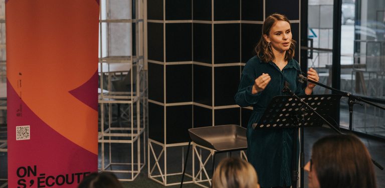 Femme aux longs cheveux blonds et à la robe verte, debout sur un podium et s'adressant à un public dans un petit auditorium.