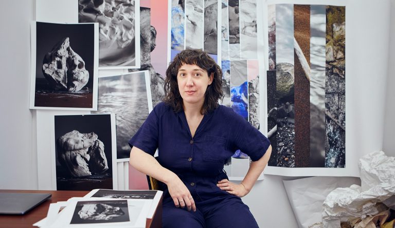 Jeune femme aux longs cheveux noirs, vêtue d'une combinaison bleue et assise à un bureau, avec ses œuvres d'art sur le bureau et le mur derrière elle.