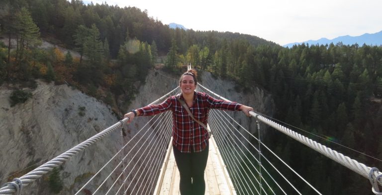 Une jeune femme souriante portant une chemise à carreaux et des jambières noires se tient sur un long pont tournant au-dessus d'un ravin.