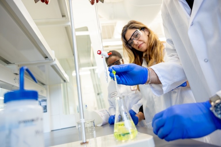 Two women look on as somebody's hands fill a beaker in the foreground
