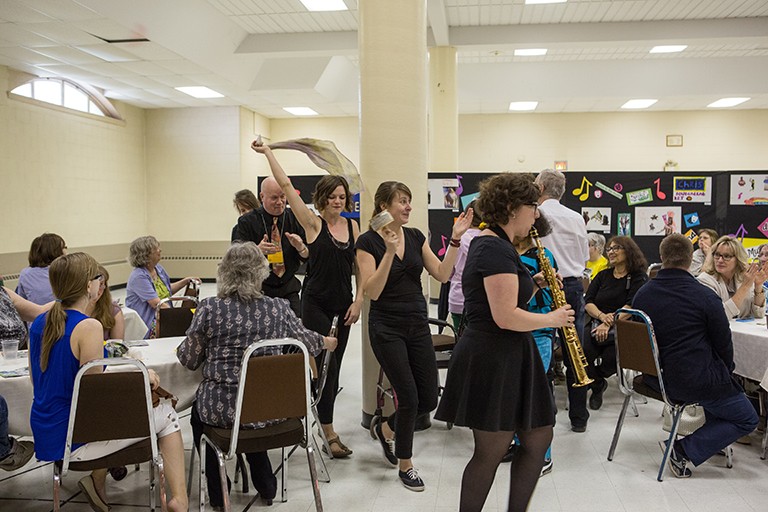 Musicians and actors dressed in black making their way through tables of seated spectators