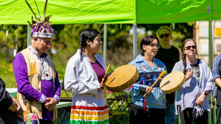 Des personnes se tenant dehors par une journée ensoleillée, vêtues de costumes traditionnels, célébrant l'inauguration d'un jardin communautaire au son des tambours et des chants.