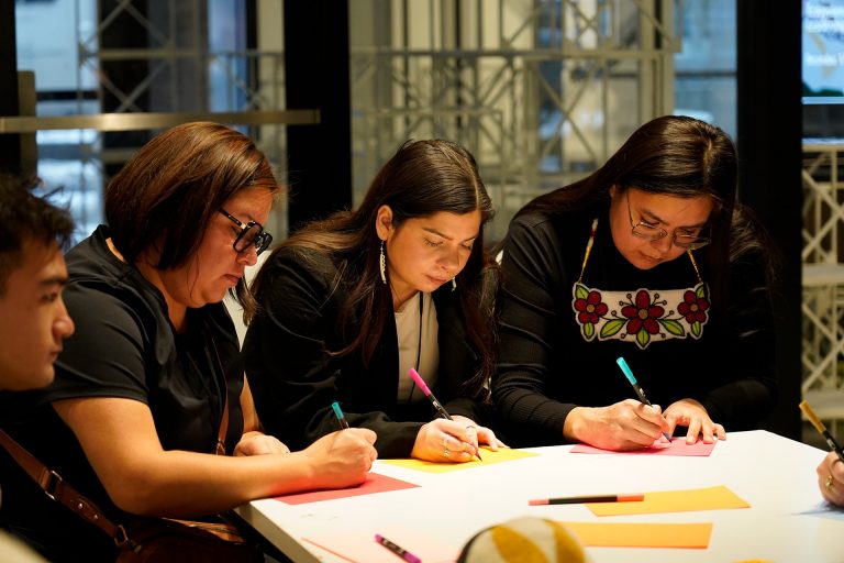 Three women sitting at a table, each with a pen and writing on coloured paper