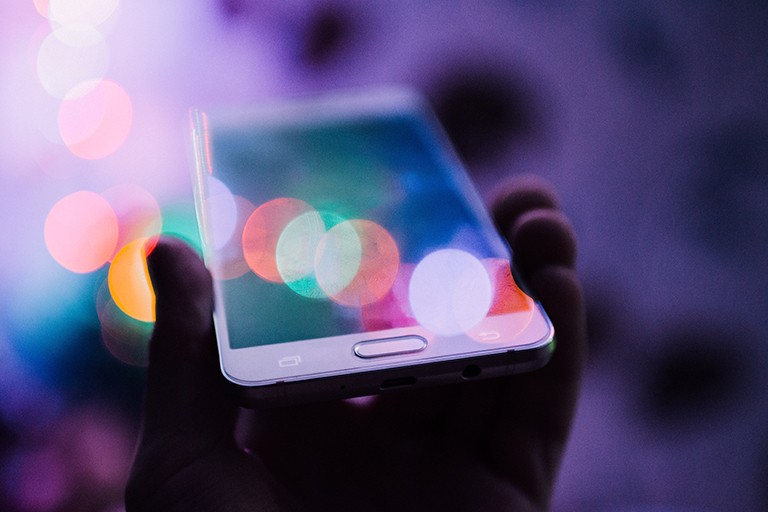 A hand holding a white mobile phone. The image is blurry with small circles of coloured light reflections. The background is light to dark purple.