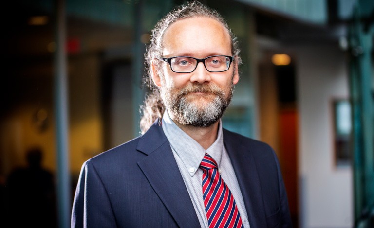 Alex De Visscher A man with long, curly hair tied back in a pony tail, with a full beard, and glasses.