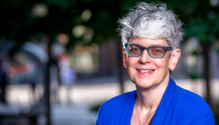 Smiling woman with short greying hair, red lipstick and glasses, wearing a blue shirt. 