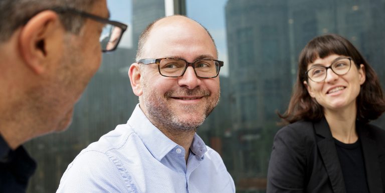 Two men and a women, wearing glasses and smiling, in a discussion with one another.
