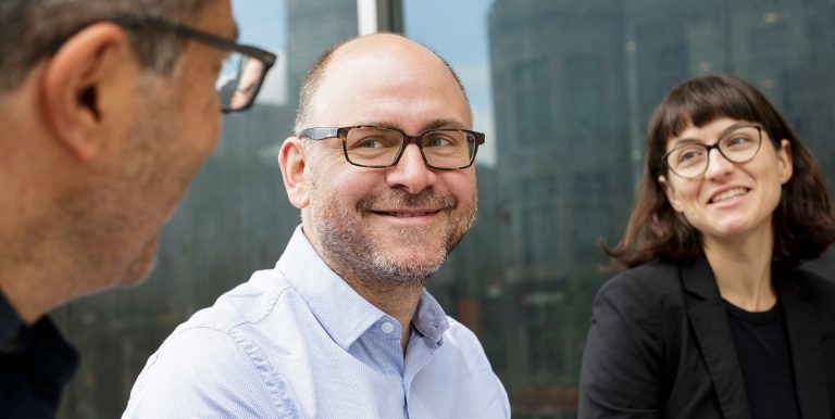 Two men and a women, wearing glasses and smiling, in a discussion with one another. 