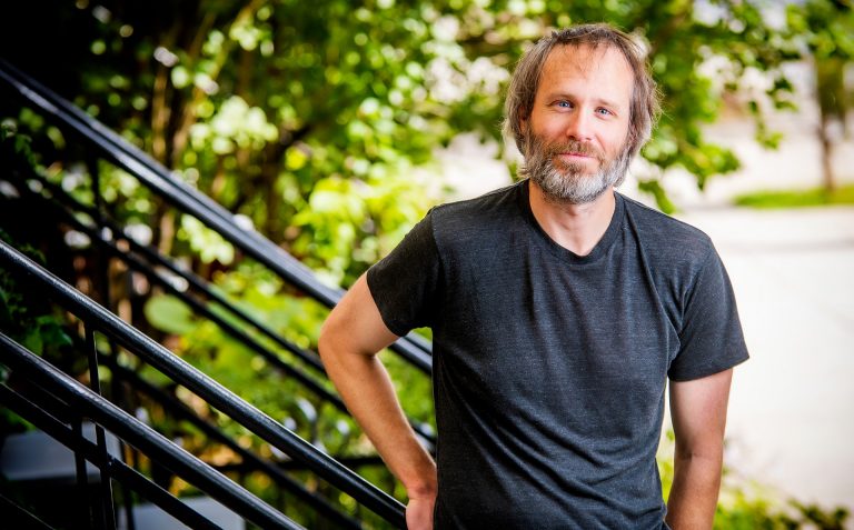 Man standing on a staircase in the outdoors, dressed in a black t-shirt, with dark hair and a greying beard.