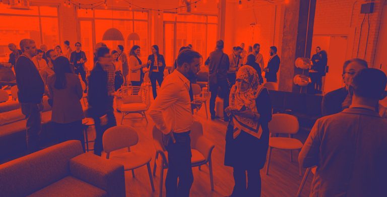 A group of people standing in a room and networking, overlaid with a red colour wash.