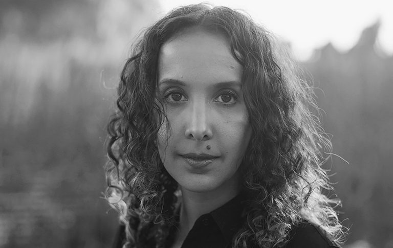 A black and white image of a young woman with long dark, wavy hair and a black shirt.