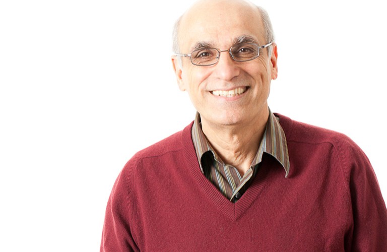 An older, smiling man with spectacles and a red knit sweater.