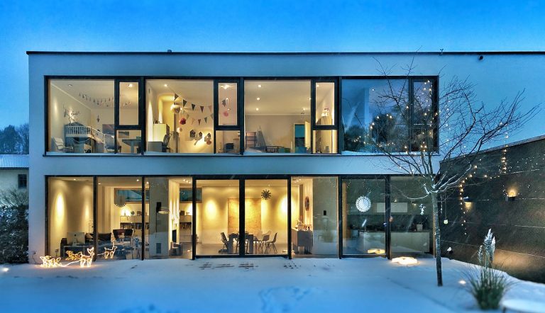 A winter photo of a residential building with large windows taken from the exterior, looking into the brightly-lit interior.