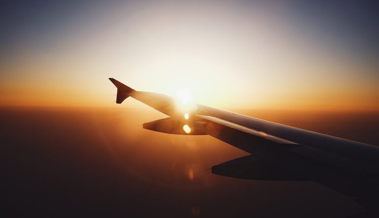 The wing of an aeroplane in flight, above the clouds, with the sunrise in the far distance.