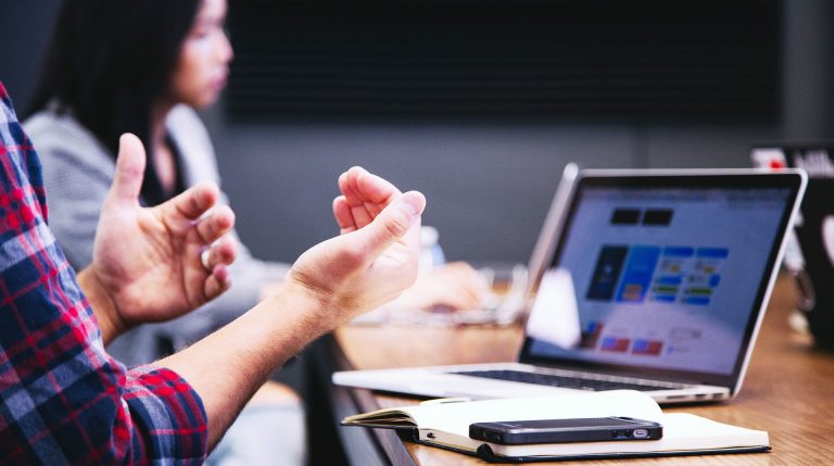 Close up of a man's hands as he talks, with a woman looking at a computer in the background.