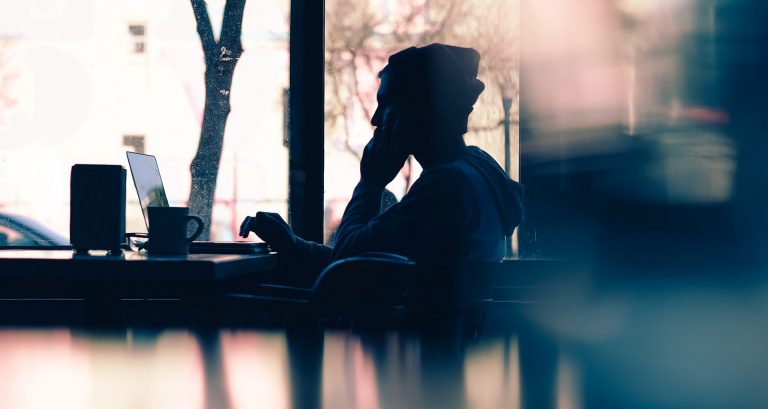 Young person, mostly in silhouette, sitting in a cafe setting and working on a laptop.
