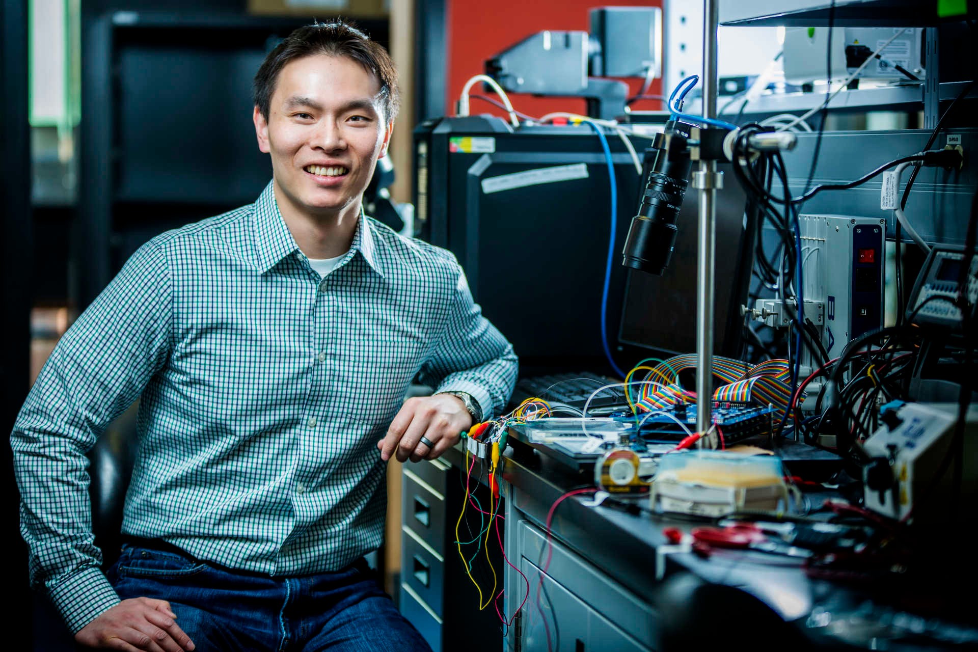 Steve Shih, a professor in Concordia’s Department of Electrical and Computer Engineering Young smiling man wearing jeans and a checked shirt, sitting in a lab with various wires and machines.
