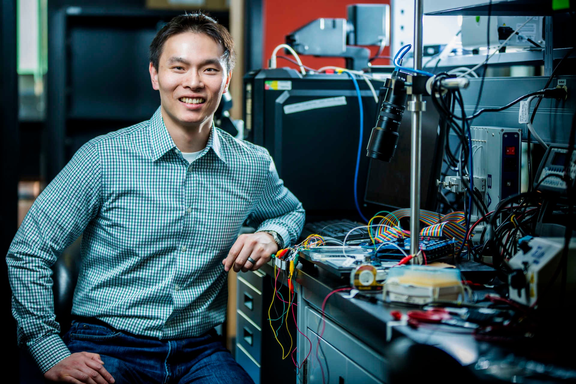 Young smiling man wearing jeans and a checked shirt, sitting in a lab with various wires and machines.