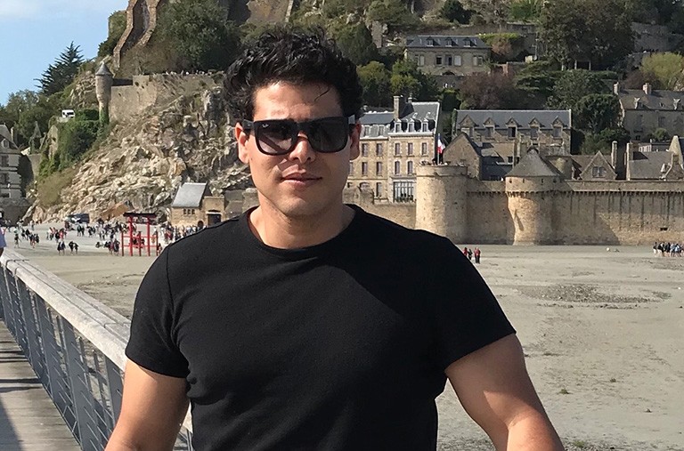 Young man with sunglasses and a black T-shirt, on a beach in a European location.