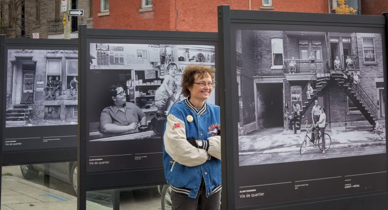 Middle-aged woman in a varsity jacket and commando pants stands looking at a photograph in an outdoor exhibition.
