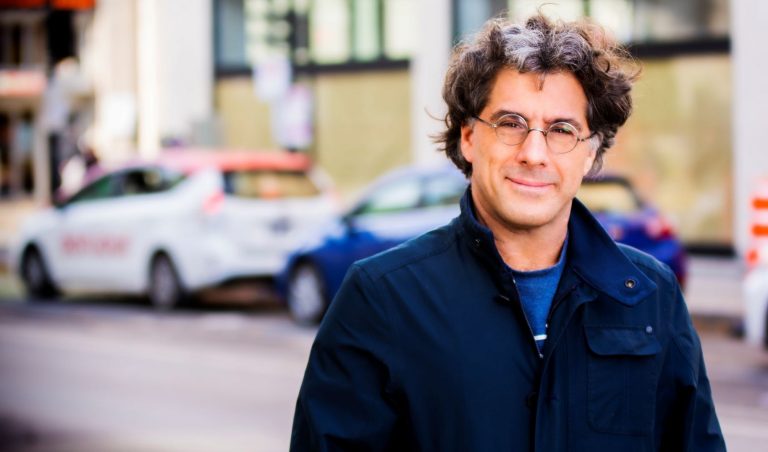 Middle aged man in glasses and blue coat standing in front of taxis