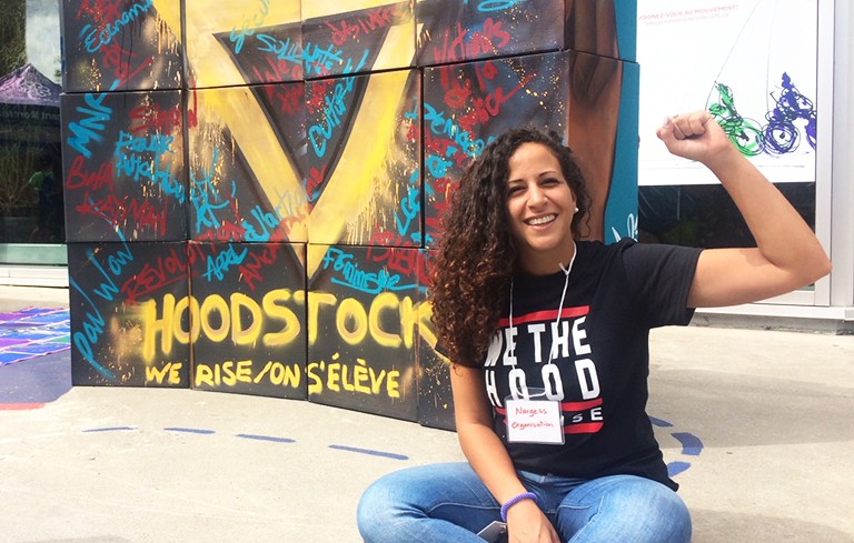 Young, smiling woman with a black t-shirt in front of a graffiti wall, on which is written "Hoodstock, we rise/on s'élève."