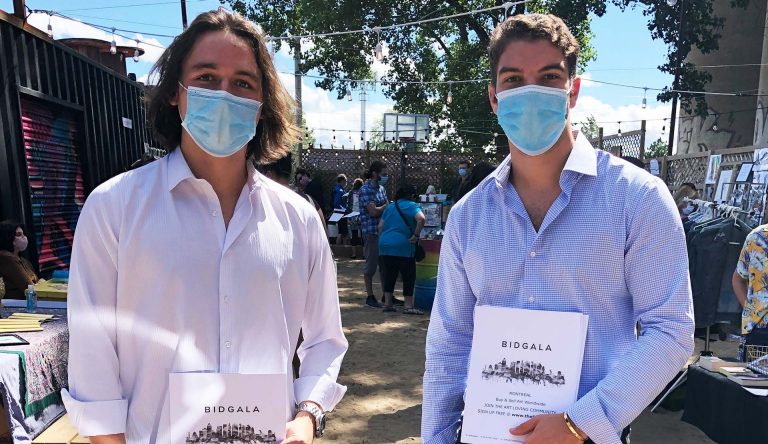 Two young men wearing masks and standing in an outdoor setting, holding booklets that say, "Bidgala" on the front.